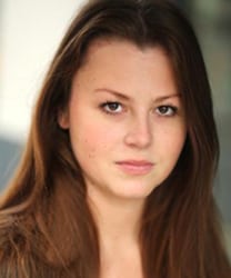 Meg Coombs smiling, wearing a blue blouse, with shoulder-length brown hair and a neutral background.