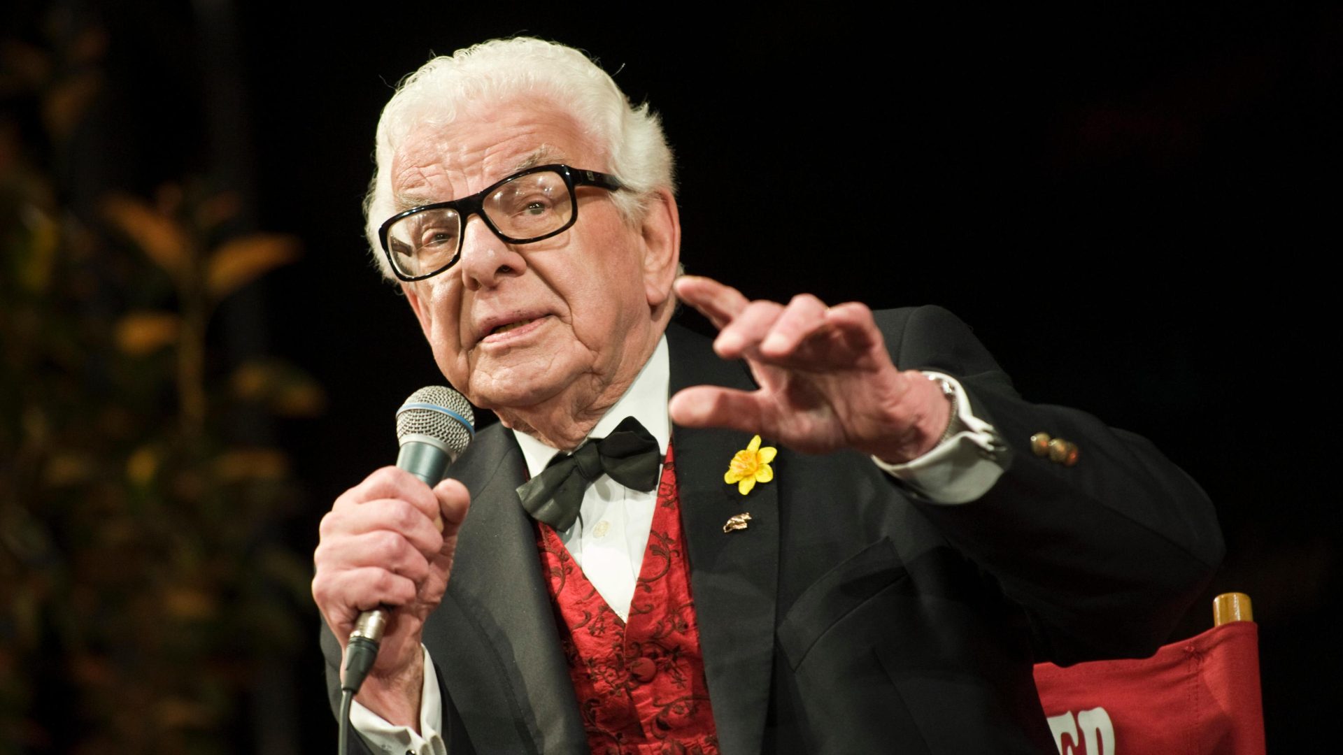 Barry Cryer smiling, wearing glasses, with a microphone in hand, on stage during a celebration event.