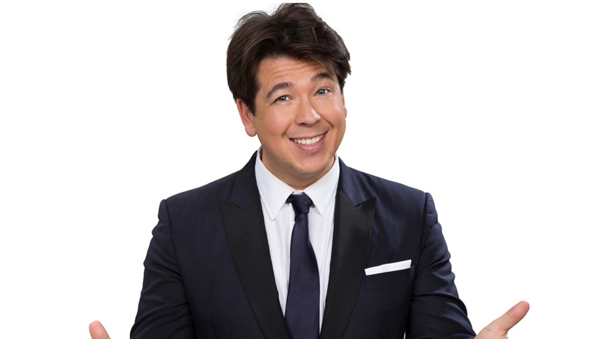 Michael McIntyre smiling, wearing a black suit, standing on stage with a microphone in hand.