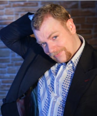 Rufus Hound smiling, with short dark hair and a blue shirt against a neutral background.