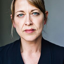 Nicola Walker smiling, wearing a black top, with soft lighting and a blurred background.