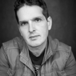 James Alper stands confidently, wearing a stylish jacket against a blurred urban background.