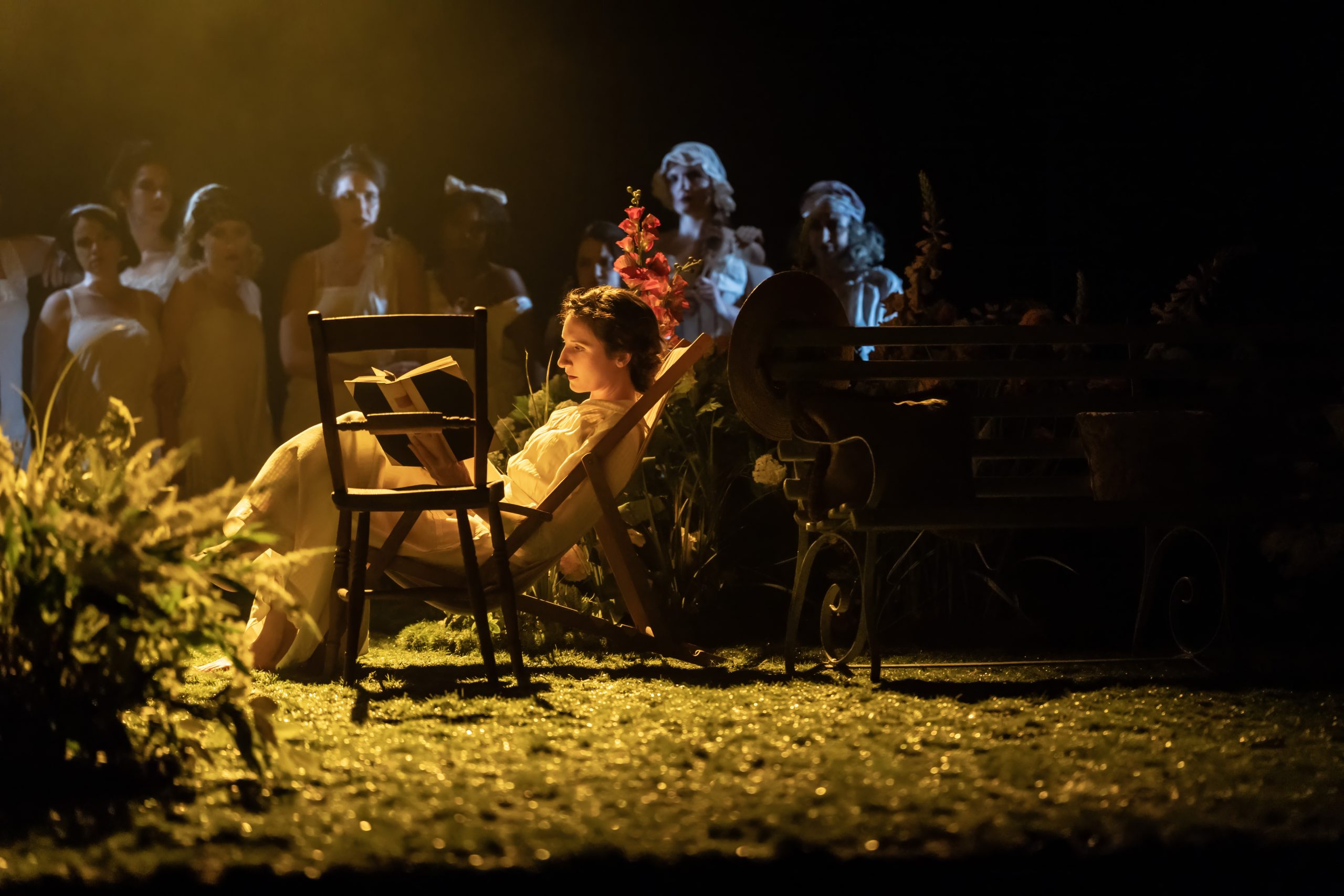 A theatrical scene featuring two women in period costumes, set against a dimly lit backdrop.