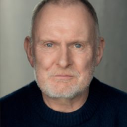 Robert Glenister in a theatrical costume, standing on stage with dramatic lighting.