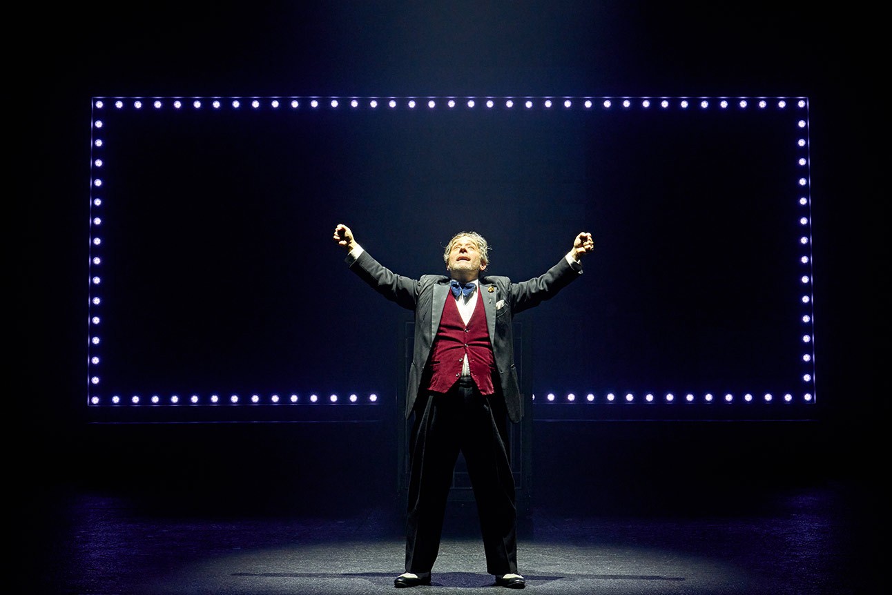 Andy Nyman as Max Bialystock, wearing a suit, smiling confidently on stage.