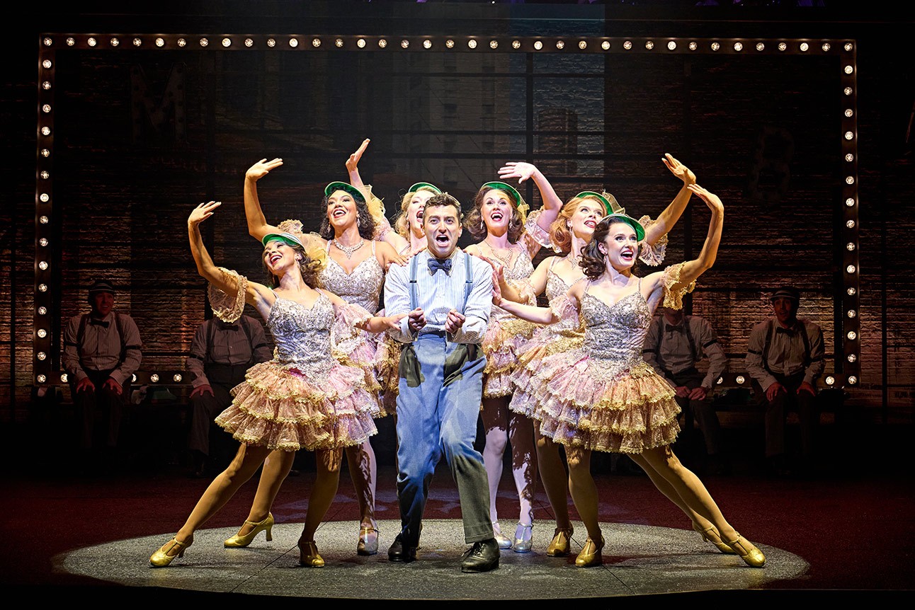 Marc Antolin as Leo Bloom with a group of performers in vibrant costumes from The Producers.
