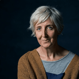 Julie Hesmondhalgh smiling, wearing a black dress, with soft lighting and a neutral background.