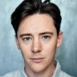 Joshua Glenister standing confidently with arms crossed, wearing a smart casual outfit against a neutral background.
