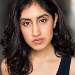Ambika Mod with a warm smile, wearing a black top, against a neutral background.