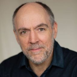 Ian Drysdale in a formal suit, standing against a textured wall, looking directly at the camera.