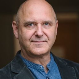 John Ramm in a formal suit, standing against a neutral background, looking directly at the camera.