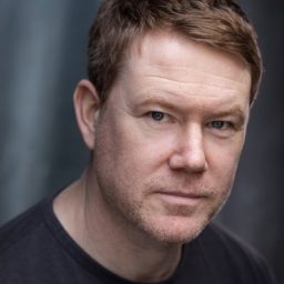 Mat Betteridge standing in a dimly lit room, wearing a dark suit and tie.