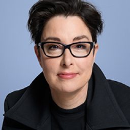Sue Perkins smiling, wearing a black top, with short dark hair and a neutral background.