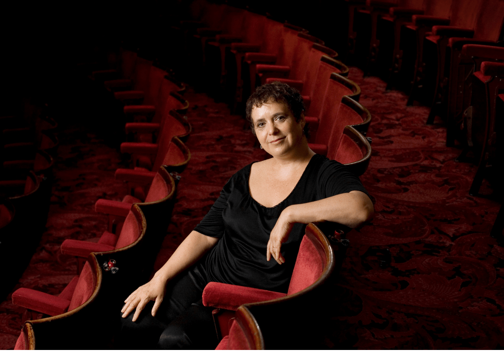 Nica standing on stage in a theatre, spotlight illuminating her as she engages with the audience.