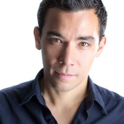 Conrad Ricamora smiling on stage, dressed in formal attire, with a spotlight illuminating him.