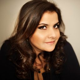 Nina Wadia smiling on stage, wearing a stylish outfit, with a spotlight illuminating her.