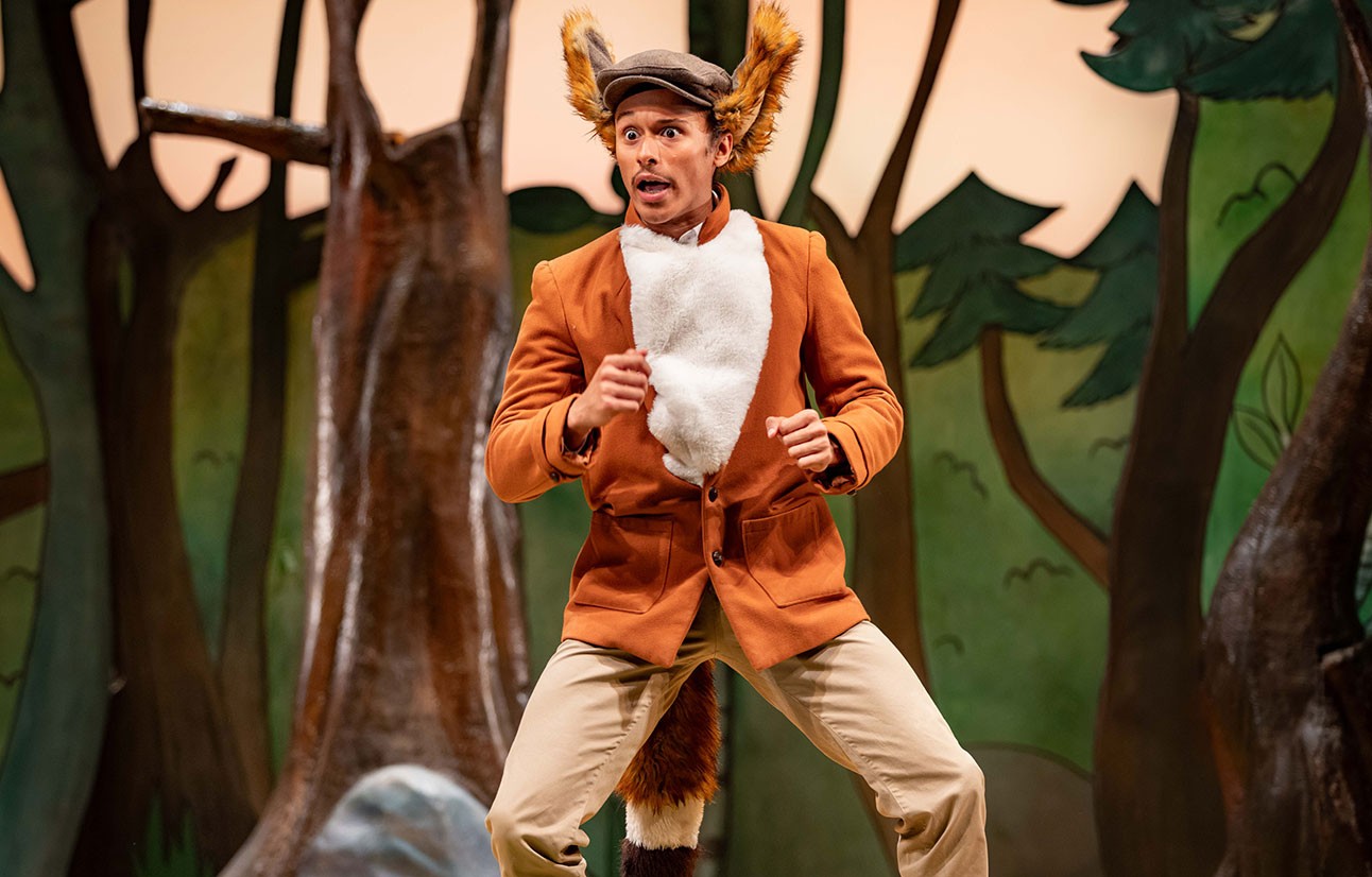 Actor in a brown fox costume with white chest fur performs on stage with forest-themed backdrop.