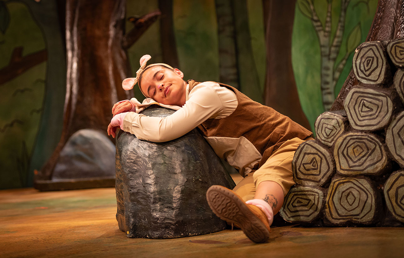 Actor in brown Gruffalo costume resting head on a grey tree stump prop on stage with forest backdrop.