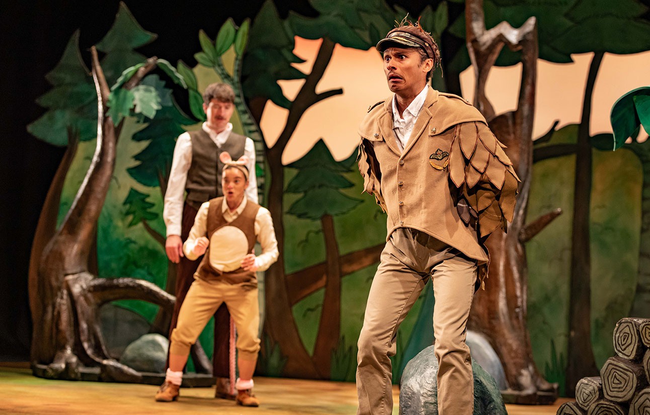 Three actors in costume perform on stage with painted forest backdrop, one dressed as an owl with wings outstretched.