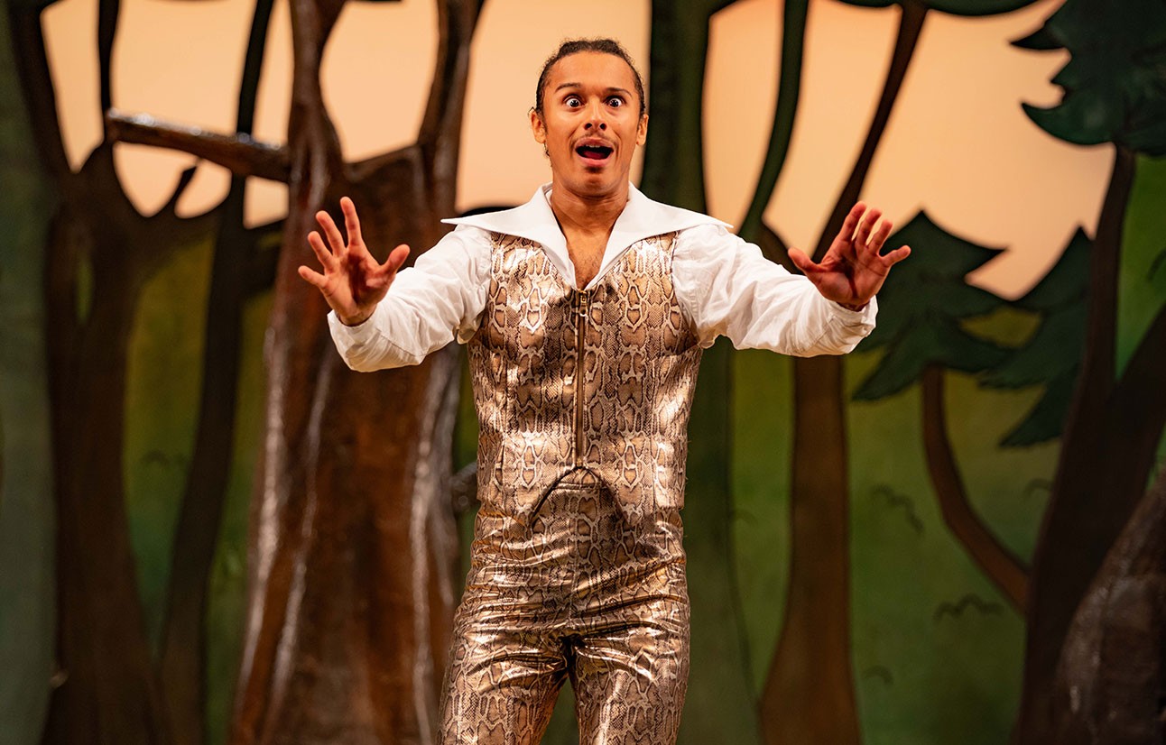 Actor in silver costume with white shirt gestures expressively on stage with stylised tree backdrop in warm lighting.