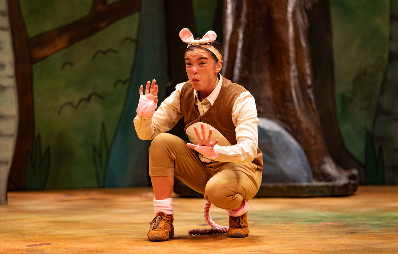 Actor in mouse costume crouches on stage with hand raised, forest backdrop visible behind.