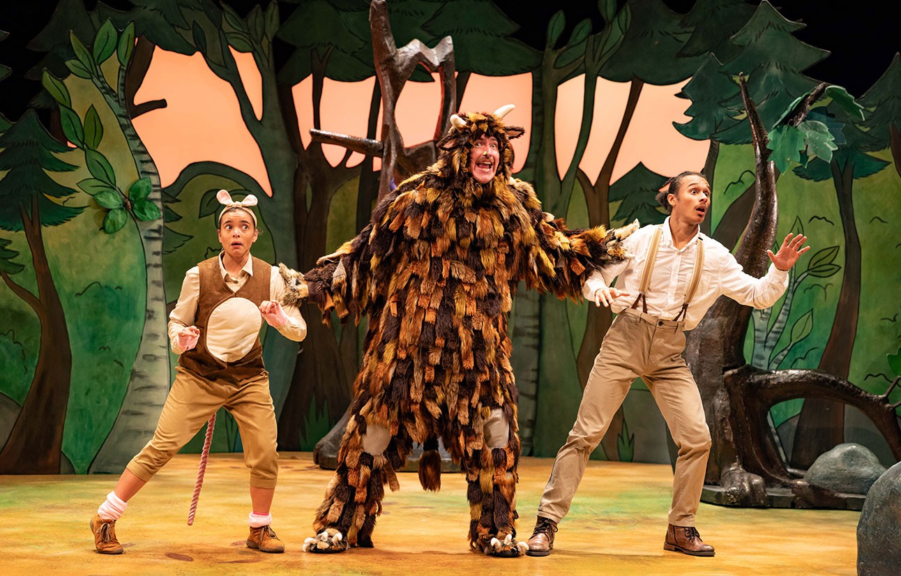 Three actors in costume perform on stage; central figure wears a brown, furry Gruffalo suit with trees in background.