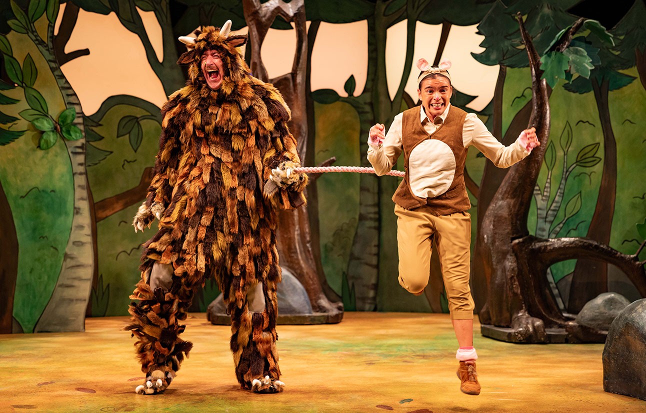 Actor in a brown leaf-covered Gruffalo costume stands beside a man in beige vest and trousers on stage.