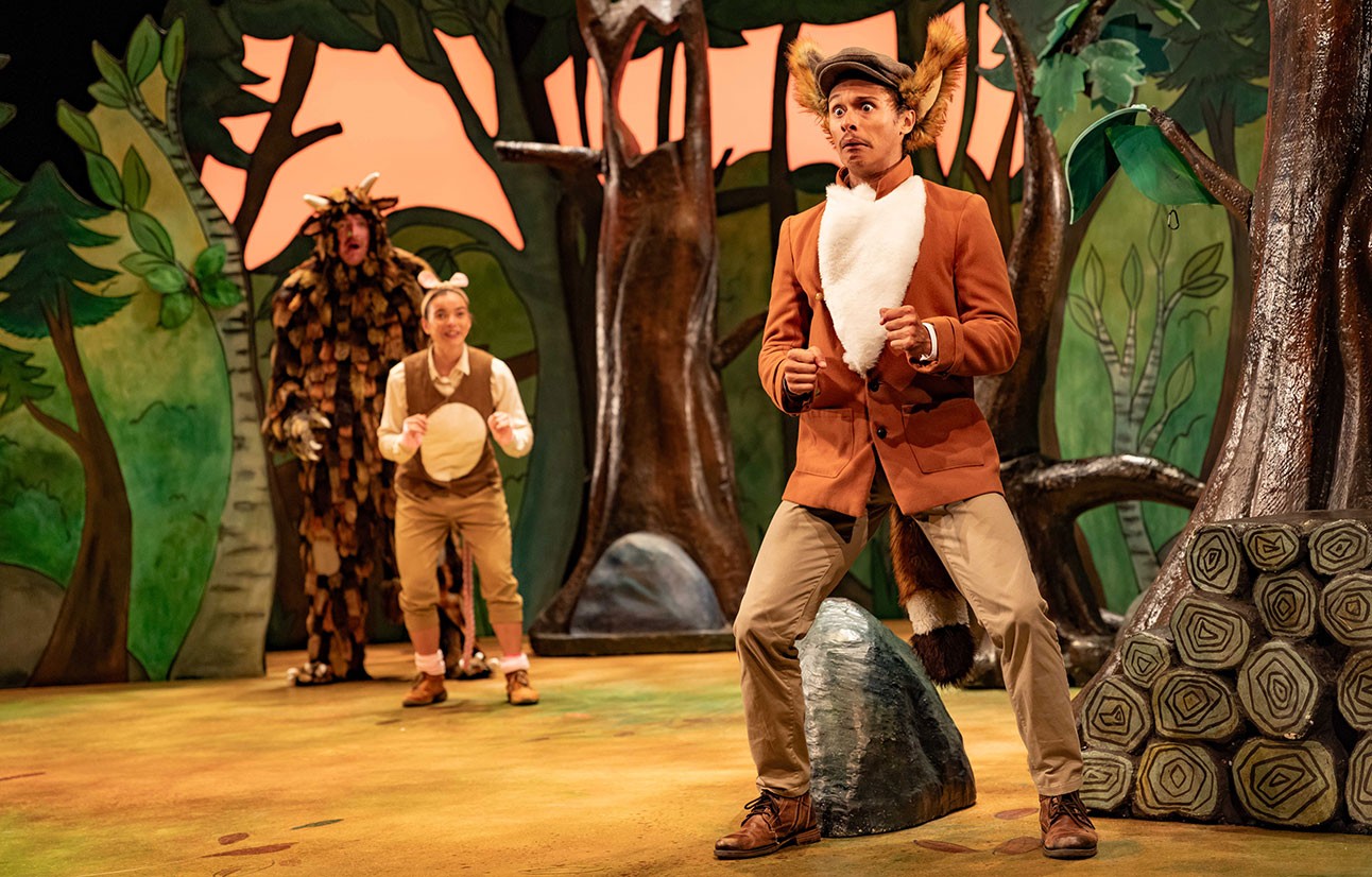 Actors in costume perform on stage with painted forest backdrop, featuring a large brown creature and two children.