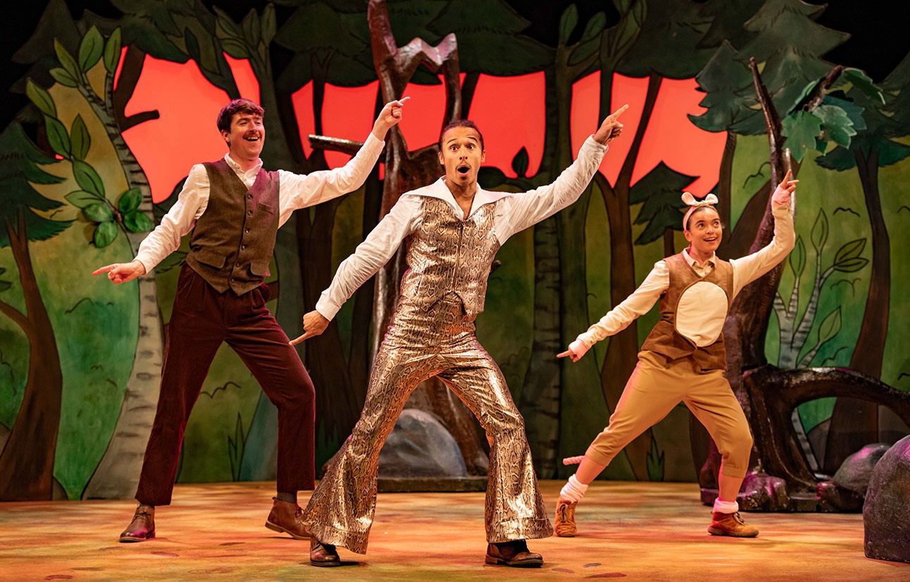 Three actors in colourful costumes perform energetically on stage with a forest backdrop and red sky.