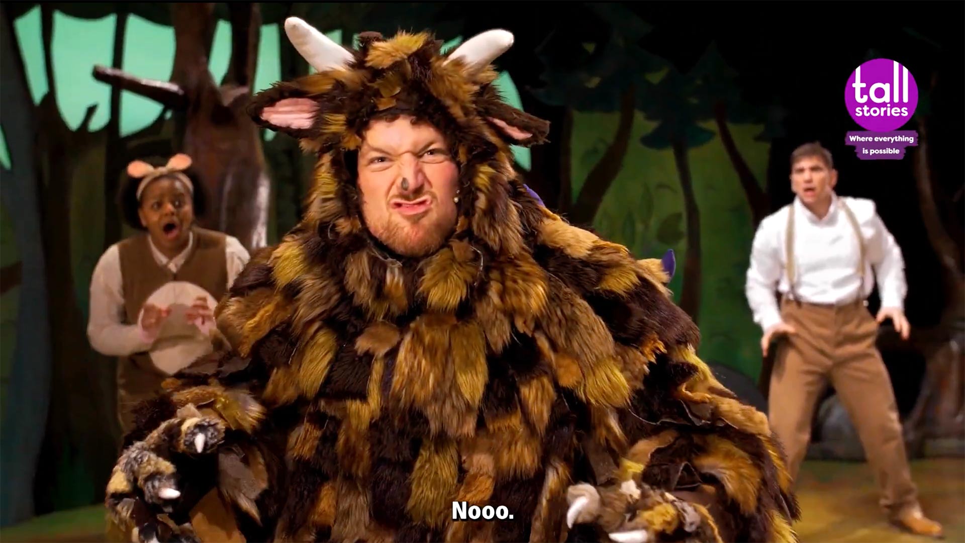 The Gruffalo character with orange eyes and purple spikes stands in a dark forest with green foliage.