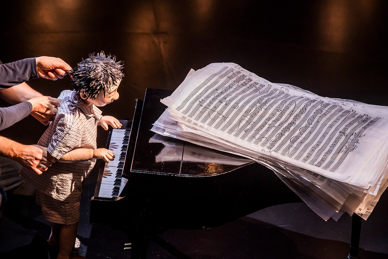 A person with spiky hair plays a grand piano with oversized sheet music on stage.