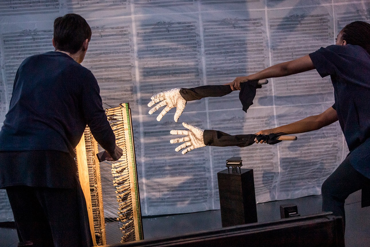 Two performers with extended arms and gloves interact with a glowing frame, set against a textured backdrop.