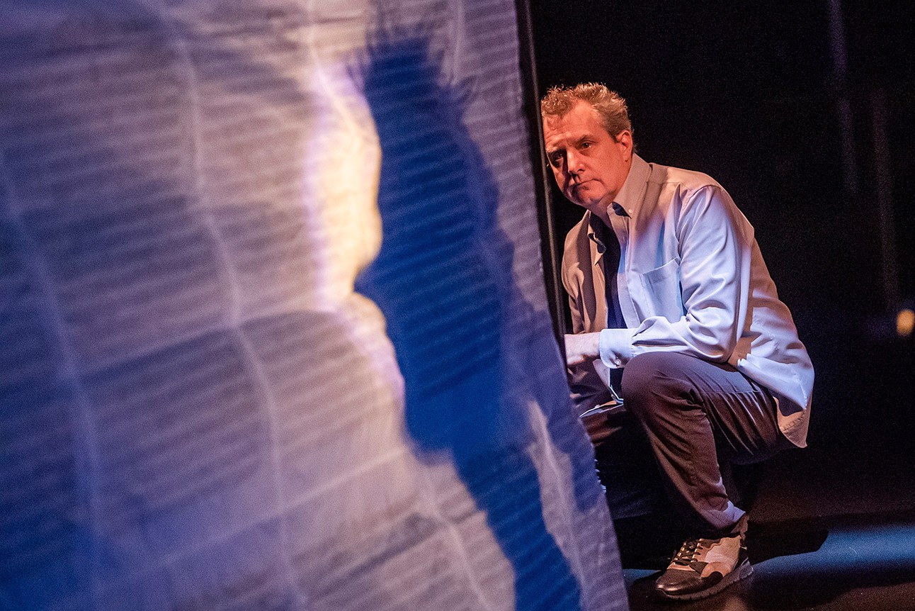 Phelim McDermott crouches beside a textured, illuminated panel casting a shadow, wearing a light shirt.
