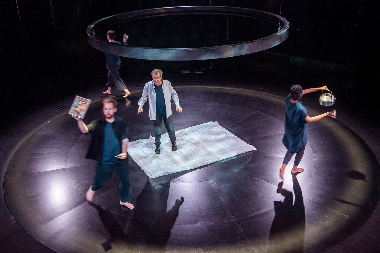 Four performers on stage, including Phelim McDermott, under circular lighting, with a central rug.