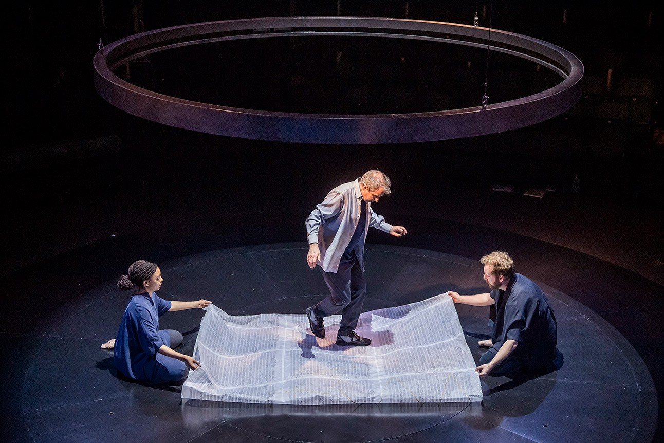 Phelim McDermott, Janet Etuk, and David Emmings perform on stage with a translucent sheet, circular backdrop.