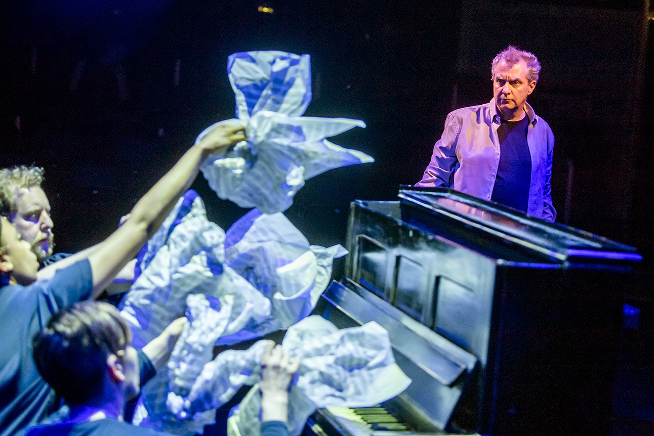 Phelim McDermott at piano, surrounded by performers holding crumpled paper, blue lighting.