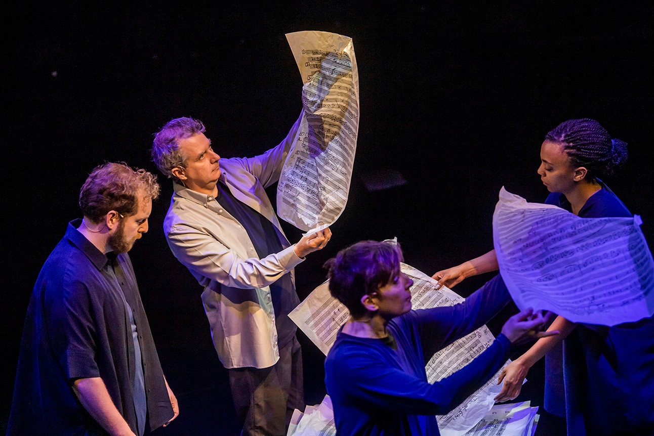 Phelim McDermott and others holding large music sheets on stage, dimly lit background.