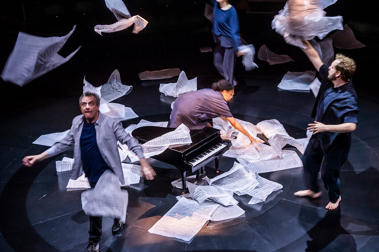 Performers on stage with scattered white sheets and a piano, wearing casual attire, dynamic movement.