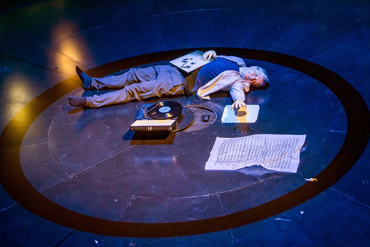 A person lies on a dark stage surrounded by papers, a vinyl record, and a turntable.