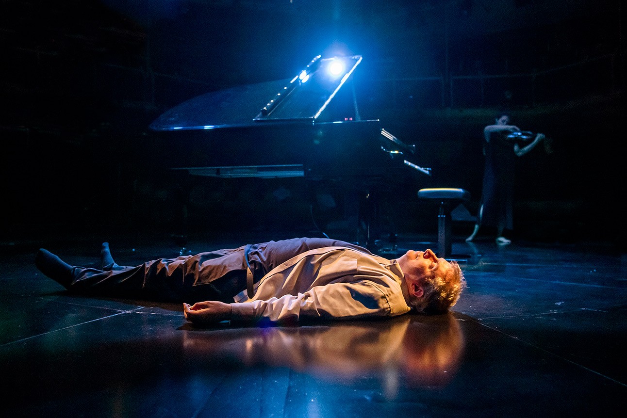 Phelim McDermott lies on stage under blue lighting, near a grand piano, during a performance.