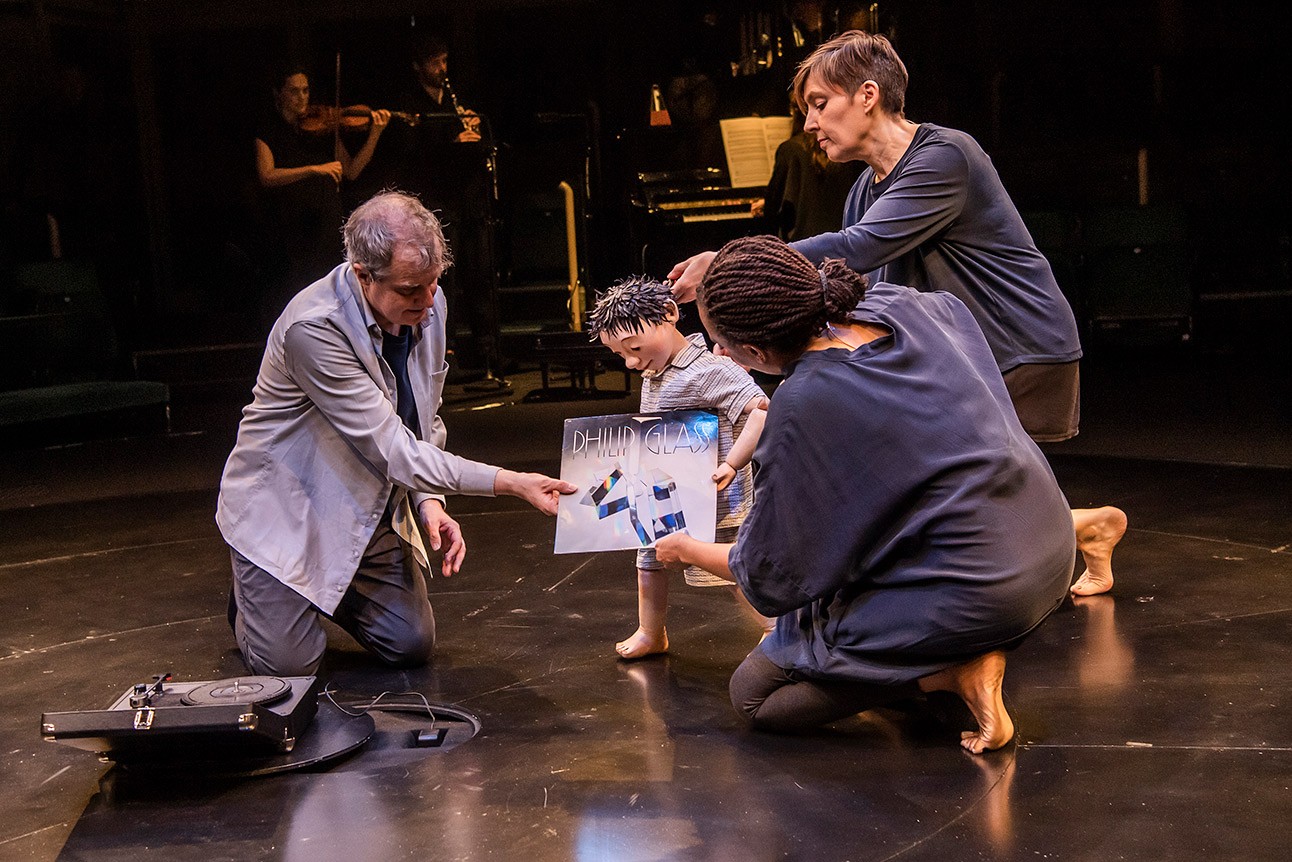Phelim McDermott, Sarah Wright, and Janet Etuk interacting with a puppet on a dimly lit stage.