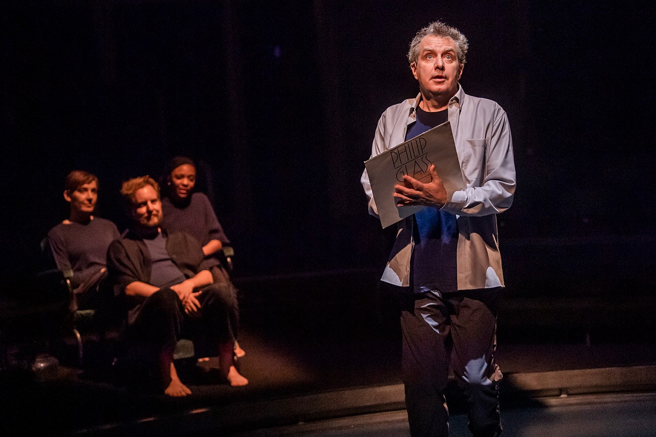 Phelim McDermott holds a book on stage, two people seated in dimly lit background.