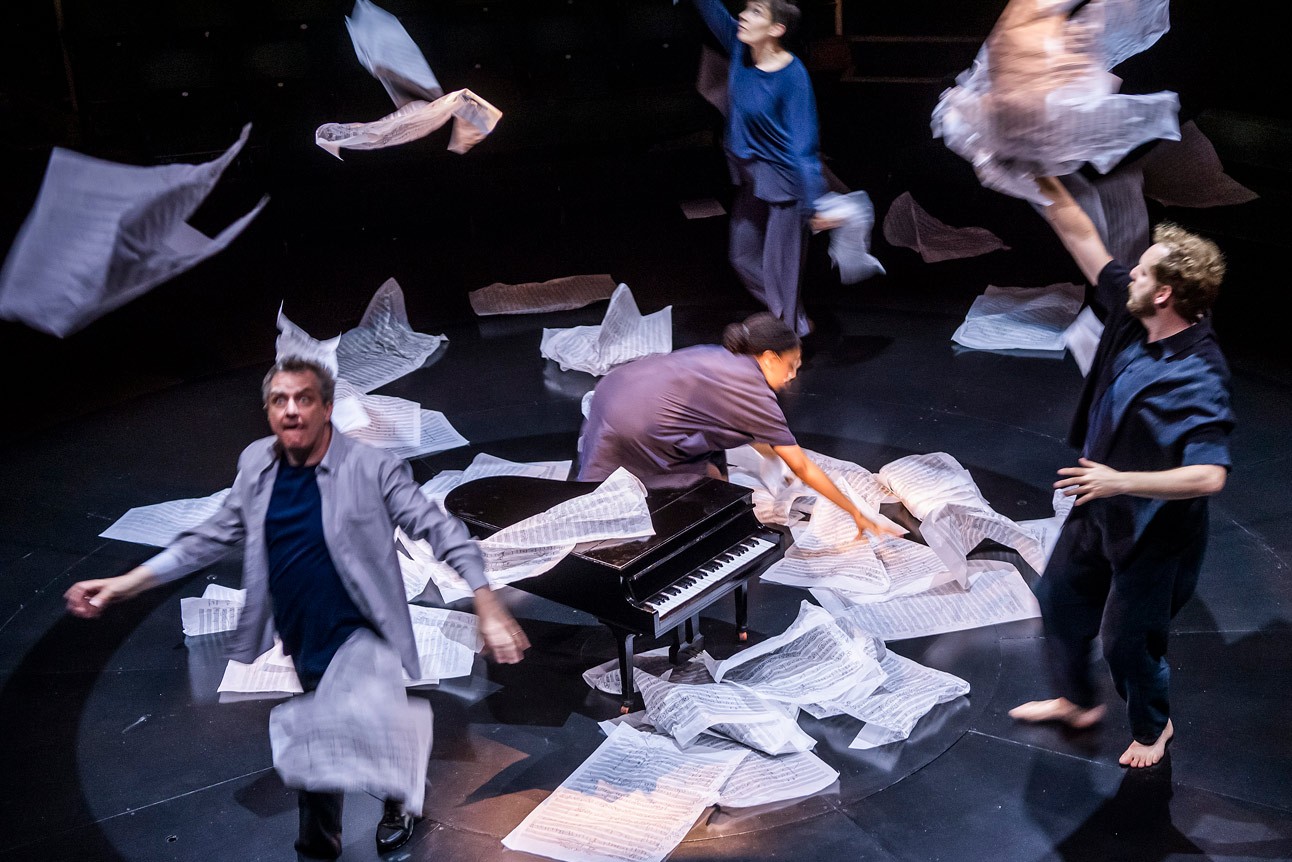 Performers energetically move around a grand piano, surrounded by scattered sheets of paper on stage.