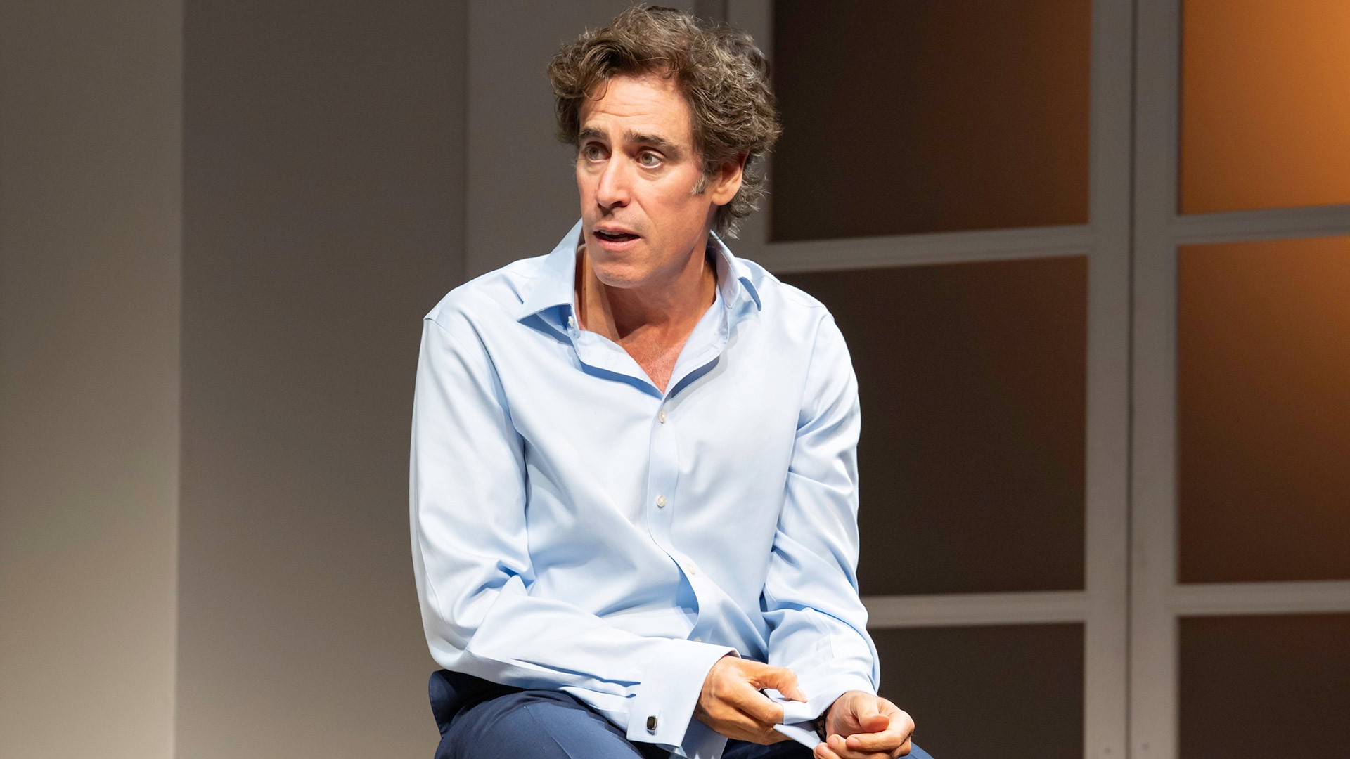 Man in light blue shirt sitting, holding paper, with a focused expression; neutral background.