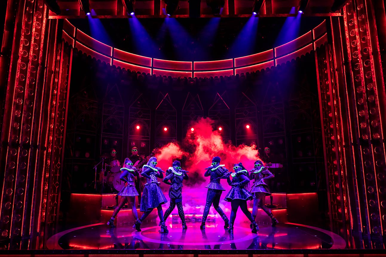Six performers in colourful costumes dance energetically on stage with vibrant red and purple lighting.