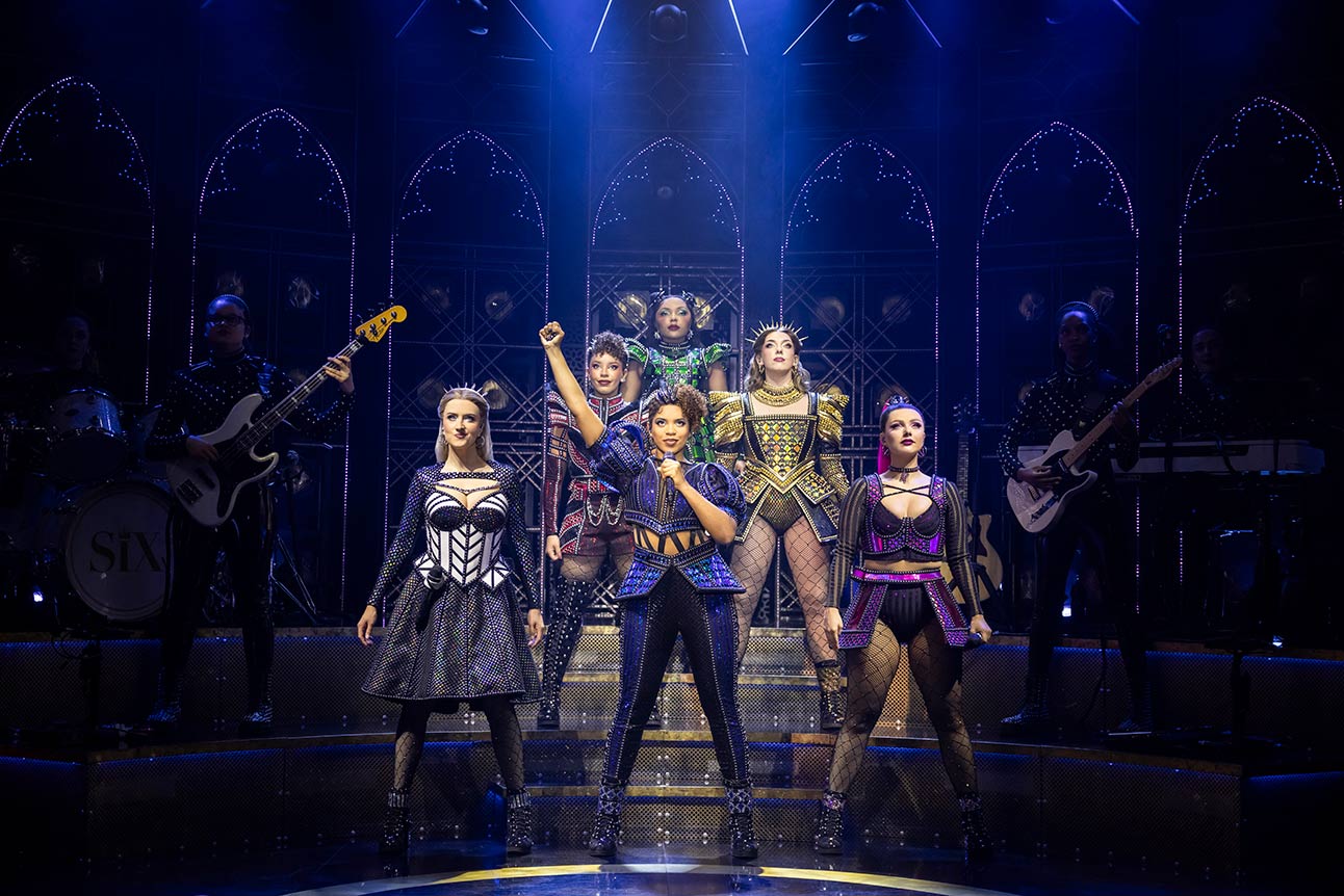 Performers in vibrant costumes sing and pose energetically on stage, with musicians playing instruments behind them.