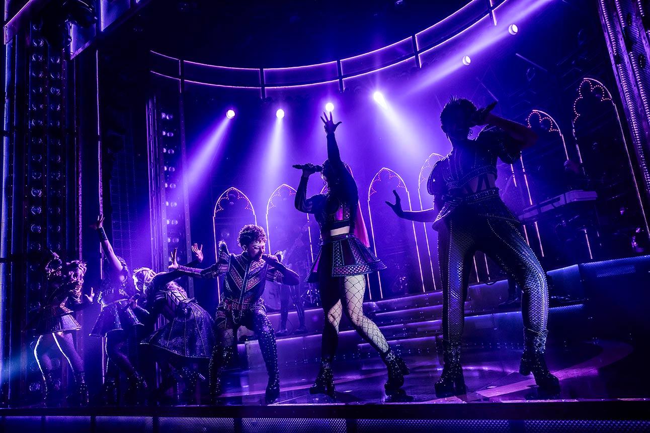 Performers on stage in dynamic poses, illuminated by purple lighting, with an arched backdrop.