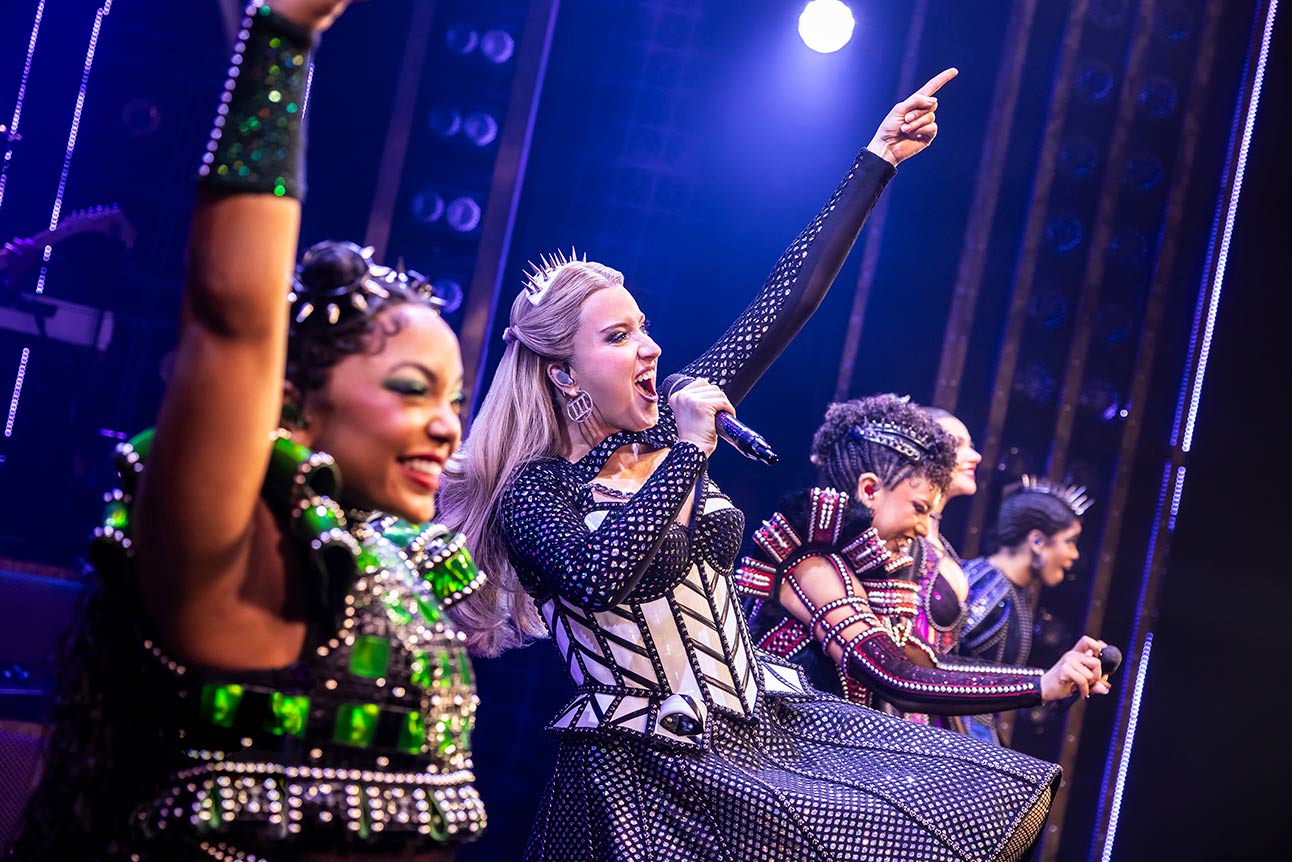 Performers in colourful costumes sing energetically on stage, with dynamic lighting and vibrant set design.