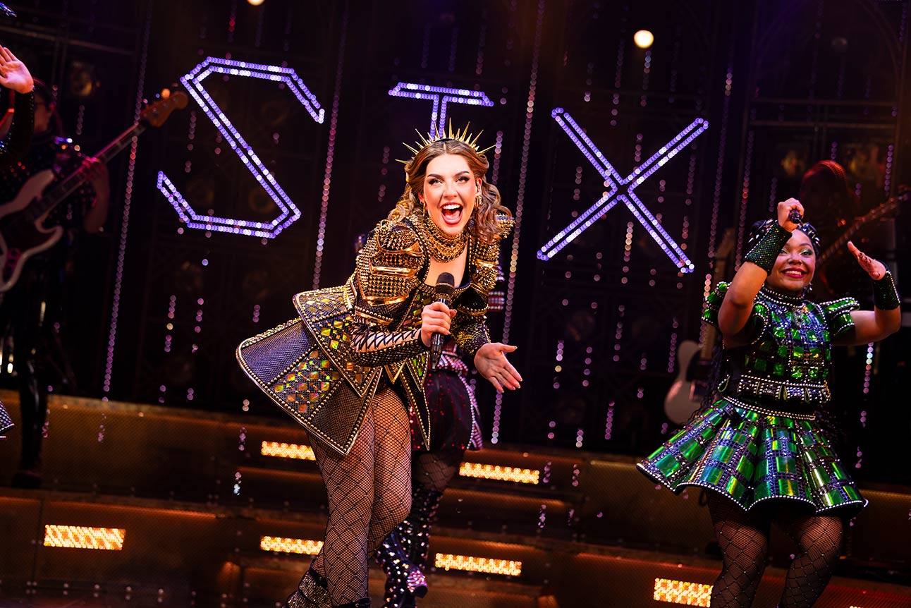 Performer in ornate costume sings energetically on stage with "SIX" illuminated in background.