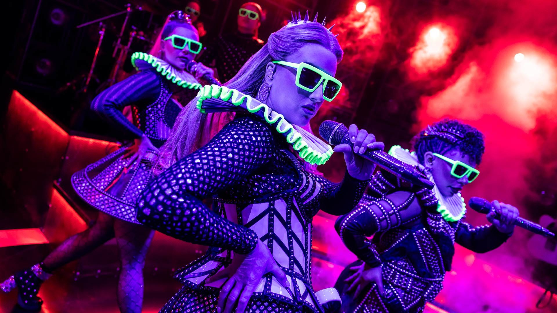 SIX Performers in futuristic costumes and neon glasses sing on stage under vibrant purple lighting.